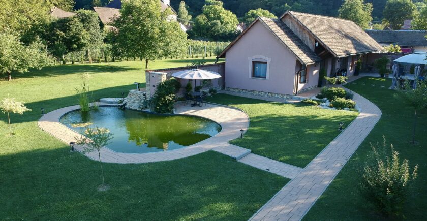 A Tulipán Vendégház épülete Kárászon, élénk zöld fűvel, sok fával, természetközeli hangulattal. Háttérben a templom torony és a Mecsek látszik. Az épület előtt a kerti tó a hangulatos kisasztallal, napernyővel ami teljessé teszi a romantikus hangulatot.