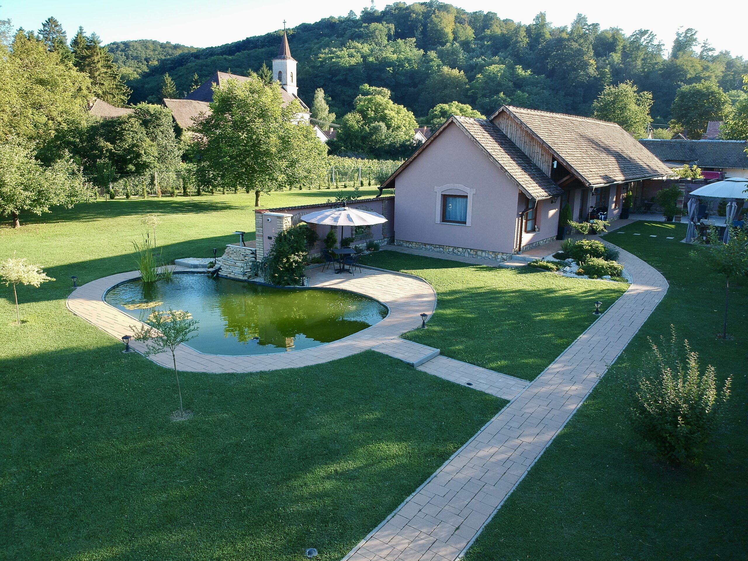 A Tulipán Vendégház épülete, élénk zöld fűvel, sok fával, természetközeli hangulattal. Háttérben a Római Katolikus templom torony és a Mecsek látszik. Az épület előtt a kerti tó a hangulatos kisasztallal, ami teljessé teszi a romantikus hangulatot.