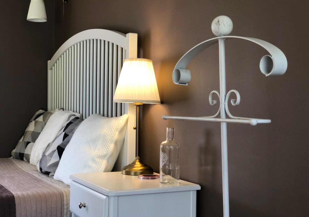 Fehér ágyvég, éjjeliszekrény és világító asztali lámpa a Tulipán Vendégház Romantik apartmanjában.