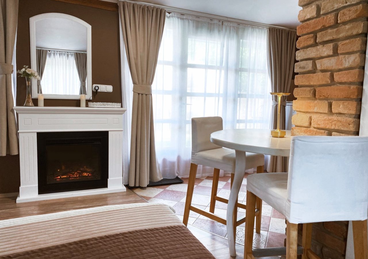 A Romantik apartman szobabelsője fehér kandallóval, tükörrel és ablak melletti reggelizőasztallal.